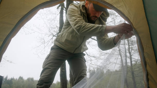 A man in a green jacket setting up a tent in a forested area. Trees and a misty, overcast sky, highlighting the outdoor and adventurous nature of camping in the wilderness.
