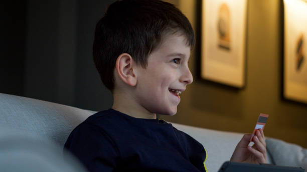 istock Young boy sitting on a sofa, engrossed in his tablet while holding a credit card, engaging in online shopping. Modern childhood, digital interaction, and tech-savvy youth in a cozy home 2165565434