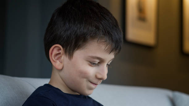 istock A young boy sits on a couch, smiling gently as he looks down in indicating a cozy home environment. 2165566083