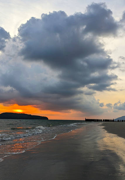 Sunset at Cenang Beach, Langkawi , Malaysia.  The hues of red light reflects on the waters and sand. The waves are reaching the shore of the beach. The sky is full of  dark nimbus clouds.