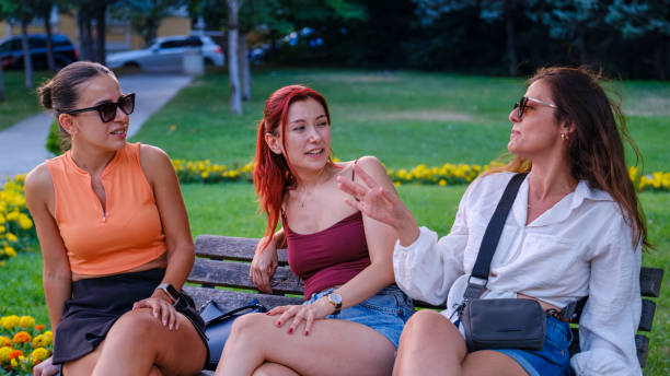 On a summer day, three beautiful young women sitting on a bench in a tree-lined park, chatting joyfully and enjoying their time together