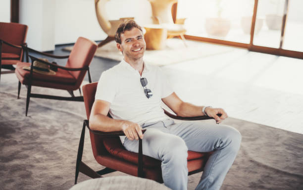 A man in a white polo shirt and light grey pants sits comfortably in a modern red armchair in a well-lit, contemporary interior space. He is smiling, wearing a wristwatch, and has sunglasses