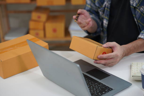 istock SME business Young man working with laptop with cardboard boxes stacked on table ready for delivery, indicating small business or home office. 2165610953