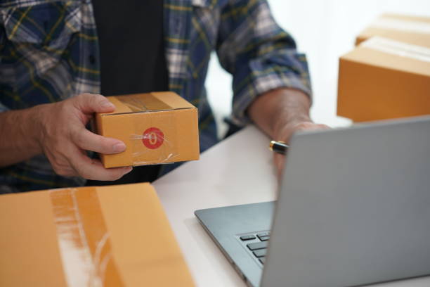 istock SME business Young man working with laptop with cardboard boxes stacked on table ready for delivery, indicating small business or home office. 2165610956