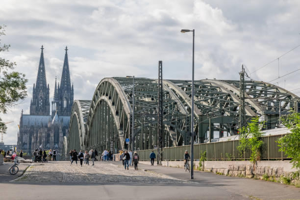 The majestic Cologne Cathedral framed by the steel arches of the Hohenzollern Bridge. This iconic view captures the blend of Gothic architecture and modern engineering in Cologne, Germany, under a cloudy sky