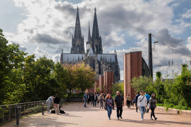 This photograph showcases the iconic Cologne Cathedral with its twin spires rising majestically above the city. The promenade in the foreground is bustling with people enjoying a pleasant day, highlighting the cathedral as a central figure in Cologne's vibrant city life.