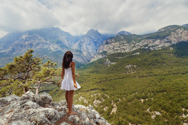 A young woman in a white dress stands on a rocky cliff, gazing at the expansive mountain landscape in Kemer, Antalya, Turkey. Perfect for travel, adventure, and nature-themed projects.