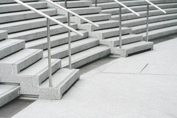 A modern staircase with wide, smooth concrete steps and sleek stainless steel handrails. The steps descend in a symmetrical pattern, creating a clean and minimalist aesthetic.