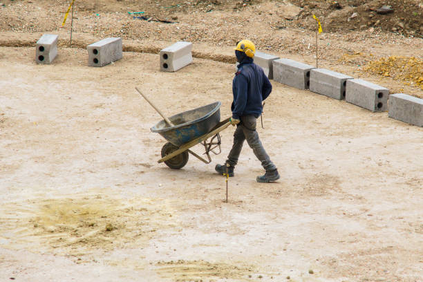 Bogota - Colombia, August 1 2024. An unrecognizable man is carrying a wheelbarrow filled with cement. The scene is set in a construction site, with a man wearing a yellow helmet and a blue jacket