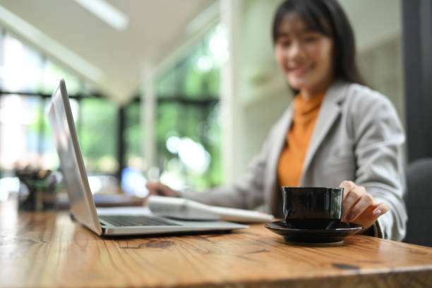 istock Smiling female office worker reaching for hot drink mug on the table while working with laptop 2165640158