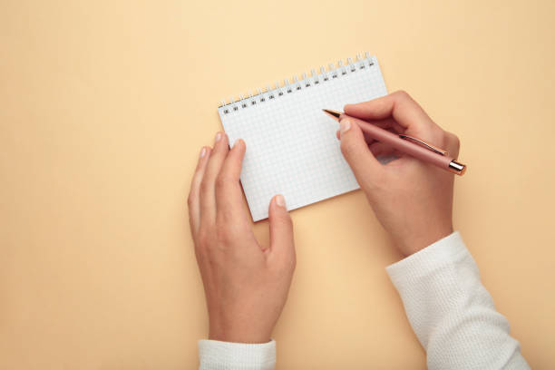 istock Woman hands hold opened notebook pages with pen on beige background. Top view 2165642495