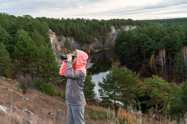 Young woman looking through binoculars while standing on a cliff near a river, Birdwatching and Ecological observations of bird migration