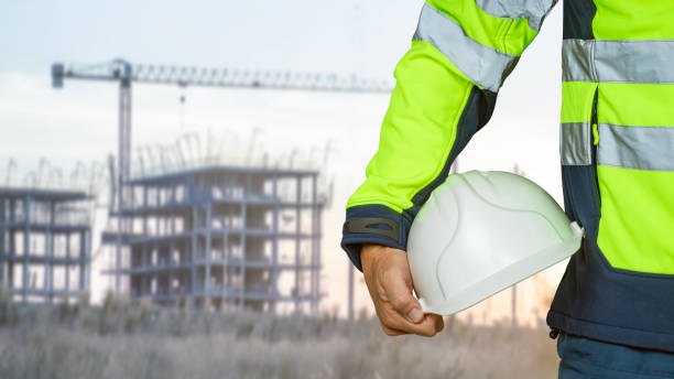 istock Construction worker in green reflective suit holding white safety helmet or hard hat with building under construction in background. 2165690125