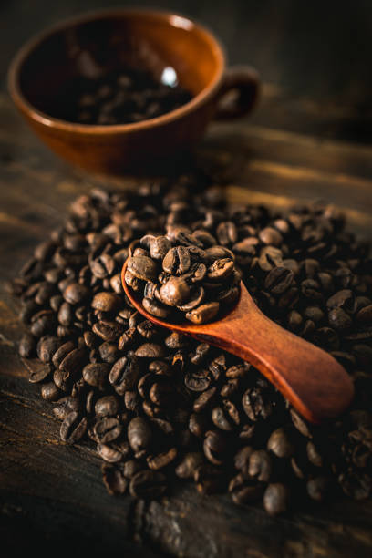 Roasted coffee beans in a wooden spoon and a coffee cup on a rustic wooden table, creating a cozy coffee scene.