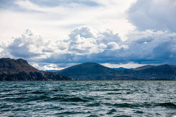 A peaceful view of Lake Titicaca from Llachon, Peru, with deep blue waters, scattered islands, and rolling hills under a bright sky with fluffy clouds