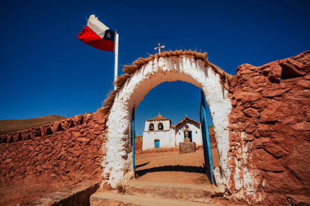 White historic church with bells, vibrant flowers, and clear blue sky in Atacama, Chile, captures culture and serenity.