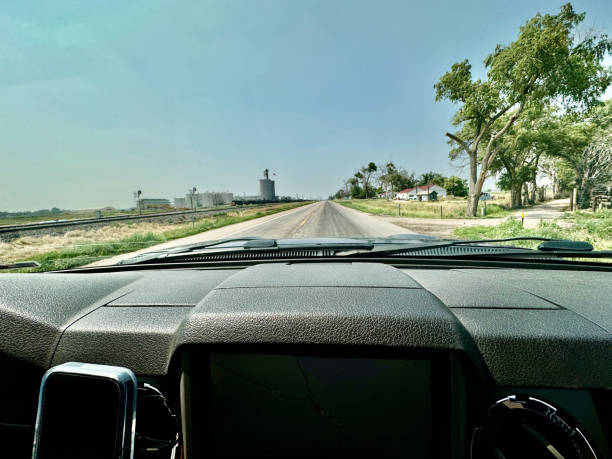 An image taken from inside a vehicle showing the dashboard in the foreground and the straight highway in the distance ahead. Railroad tracks run alongside.  A mill can be seen on one side of the road and farmhouses on the other side of the road.