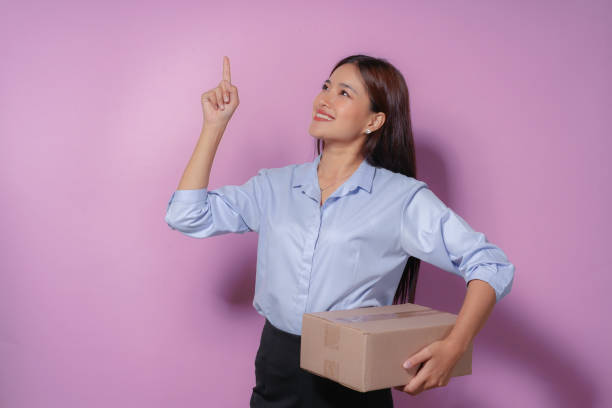 istock Young entrepreneur is holding a shipping box and pointing upwards with her index finger while smiling, suggesting a great new idea for her business 2165806730