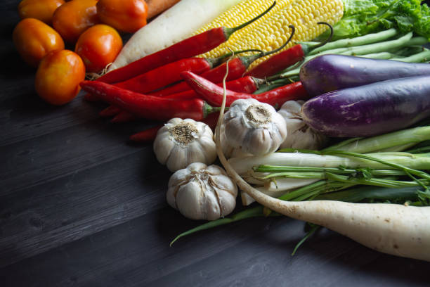 Stock image of a colorful assortment of fresh raw vegetables arranged on a rustic wooden table. Featuring corn, tomatoes, garlic, lettuce, radish, beans, chili, eggplant, carrot, bok choy, Chinese cabbage, chayote, and spring onions. Perfect for food blogs, healthy eating concepts, organic farming, and culinary visuals.
