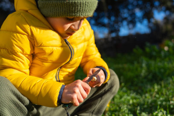 istock Boy peeling a stick with a razor in nature in warm clothes. risk play 2165818708