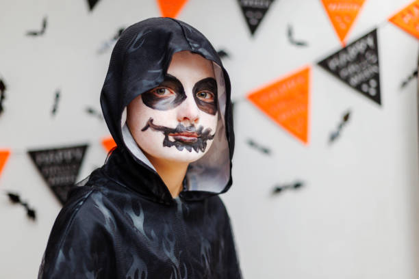 Child dressed in a black hooded costume with skull face paint, standing in front of Halloween decorations. Studio portrait with festive decor.