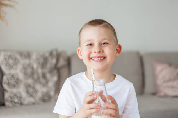 Adorable happy fair-haired boy in light clothes drinks milk from a glass bottle and eats a loaf of bread at home. Eco products. Healthy food and drinks. Milk.