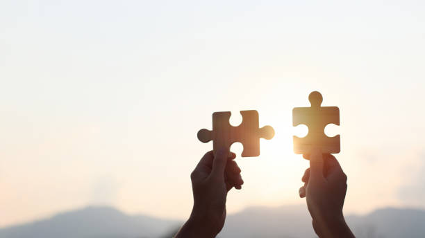 istock Teamwork and partnership concept. Hands are raised against a bright, white sky, each holding a piece of a wooden jigsaw puzzle. teamwork, problem-solving, Charity, volunteer. Unity, team business. 2165840661