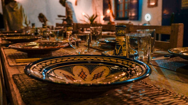 A beautifully crafted wooden table is adorned with an array of vibrant Moroccan dishes, showcasing the rich culinary heritage of the region. Each plate is intricately designed, featuring colorful patterns and textures that reflect the artistry of Moroccan craftsmanship. The table's rustic charm complements the dishes perfectly, creating an inviting atmosphere for sharing meals and stories. Aromatic spices and fresh ingredients hint at the delicious flavors awaiting to be savored, while the warm lighting casts a cozy glow over the setting. This scene captures the essence of Moroccan dining, where every meal is a celebration of culture, tradition, and togetherness.