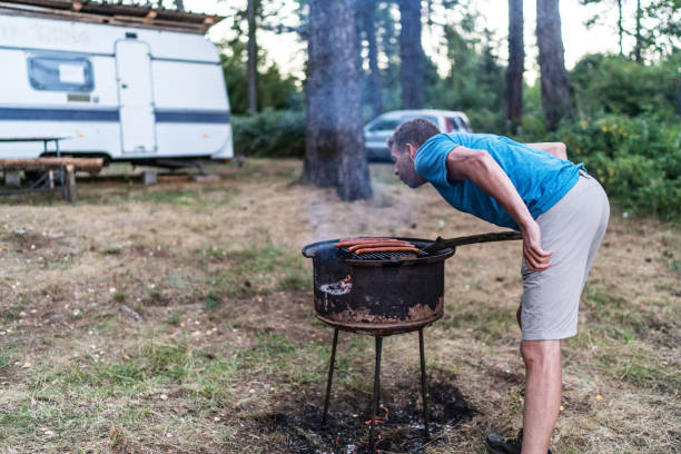 Man cooks shashlik over campfire on the lake shore, grilling meat skewers in nature for outdoor barbecue picnic on spring day, enjoying cooking food