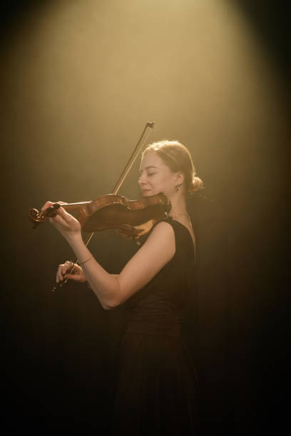 Vertical side view shot of smiling female classical music performer playing violin under projector light in warm light studio, copy space