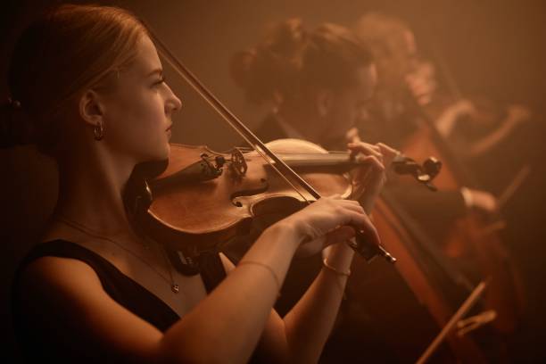 Talented Violinist Playing Music with Orchestra in Studio Side portrait of virtuoso female classic music artist playing violin with string quartet on stage in misty orange light string ensemble stock pictures, royalty-free photos & images