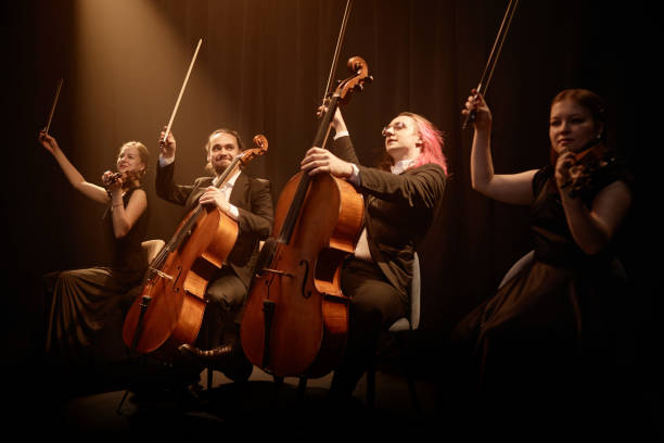 Four Professional String Players Giving Concert on Stage Chamber quartet of string musicians sitting in row on stage with project light raising bows while thanking public at end of performance string ensemble stock pictures, royalty-free photos & images