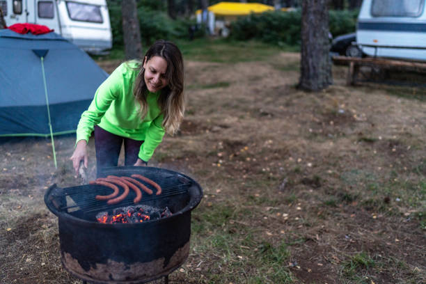 Young woman grilling sausage on skewer over campfire