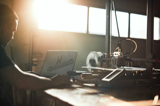 An employee designs a 3D element on a laptop that is being printed on a 3D printer in a sunlit workshop. The image captures the creative process with a striking contrast of light and shadow.