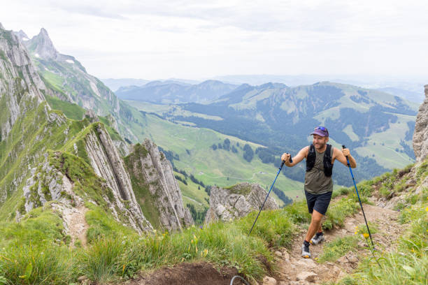 A man wearing a cap and backpack climbs a steep mountain trail with trekking poles, surrounded by rugged cliffs and scenic green valleys.