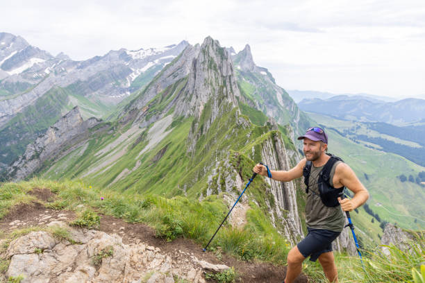 A man with a cap and backpack hikes with trekking poles on a mountain ridge, surrounded by dramatic peaks and lush green valleys.