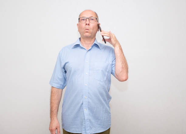 A middle-aged Caucasian man wearing glasses with a funny expression on his face speaks on his smartphone and listens attentively