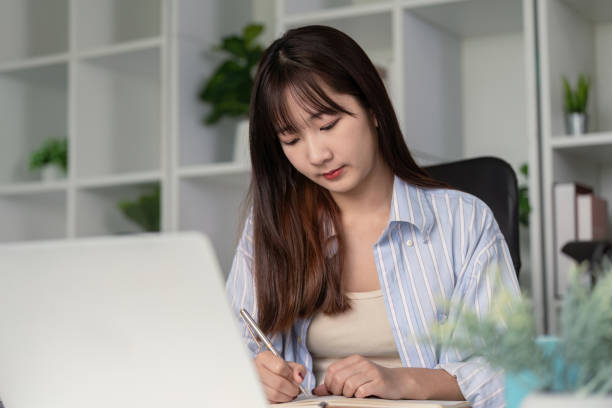istock Young Student Studying Online at Home Using Laptop and Taking Notes in a Modern Home Office Setting 2165956342