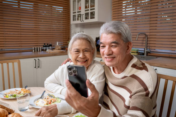 istock Asian senior couple using phone video call while having dinner in house. Attractive loving elderly mature man and woman feel happy and relax, enjoy using smartphone and eating meal in kitchen at home. 2165972810