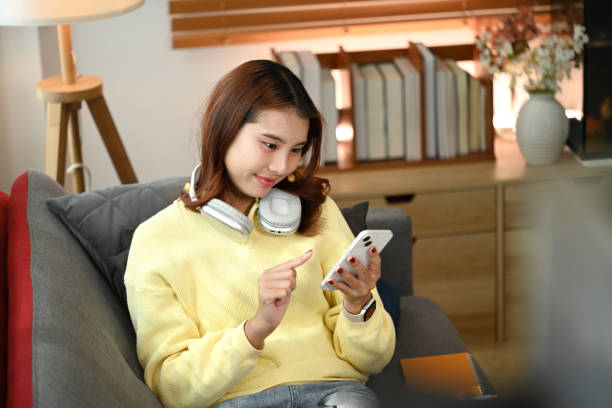 istock Relaxed asian girl with headphone around neck using mobile phone while lying on sofa at cozy living room 2165980438