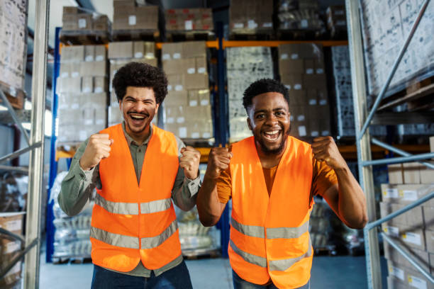 Exited multicultural warehouse workers cheering and celebrating victory at facility while smiling at camera.
