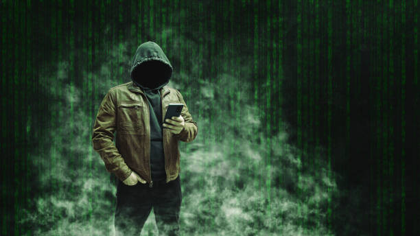 istock Faceless man, hooded cybercriminal terrorist, anonymous hacker standing and using smartphone on a dark green background with a smoke 2166028600