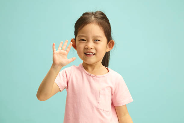 Delighted Lovely five year old kid Girl joyful waves her hand to you stands over blue isolated. Cheerful little child portrait on turquoise background. Horizontal Portrait of Sweet Asian ethnicity child girl in pink t shirt joyful waving hello, cheerful smiling to camera, has a positive mood, stands against blue isolated background with a free empty space. Happy childhood concept. Cheerful little child portrait on turquoise background. kid waves stock pictures, royalty-free photos & images