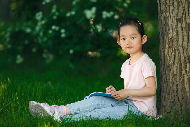 istock Young Girl Writing in a Notebook While Leaning Against a Tree in a Park 2166043573