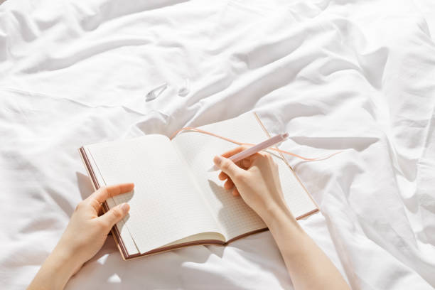 istock Woman makes plan, day results on blank white page diary journal on bed, writing in notebook or wish diary, holds notepad and pen in hands, Increasing mindfulness, relax and comfortable concept 2166053276