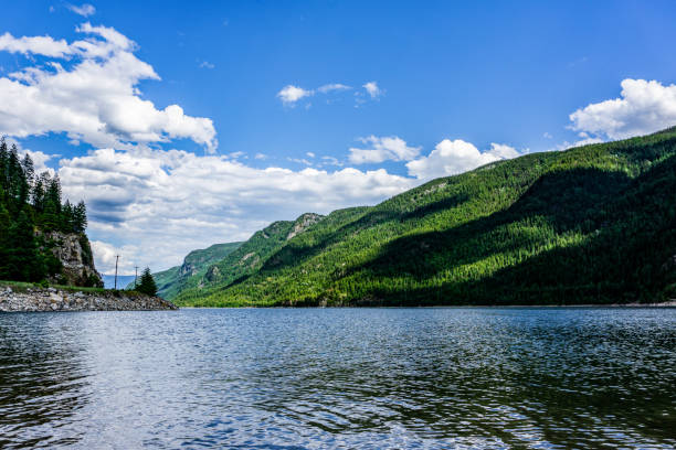 This tranquil image captures the peaceful shoreline of the Columbia River in Canada, with lush green mountains casting long shadows across the water. The river flows gently, reflecting the clear blue sky and the rich, green forest that envelops the surrounding landscape. The scene exudes a sense of calm and serenity, inviting viewers to immerse themselves in the natural beauty of the Canadian wilderness. The rugged cliffs and dense trees stand as guardians over the river, creating a perfect harmony between land and water.