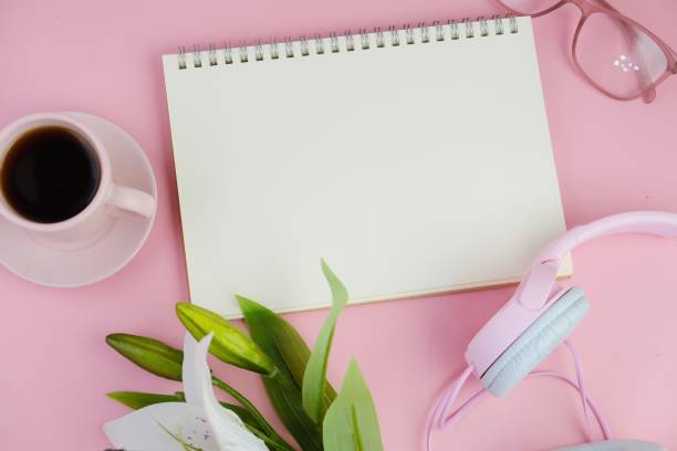 Flatlay of coffee with office tools, pink headphone, laptop, spectacles, notebook, pen and a bouquet of white daisy on pink background as copy space concept
