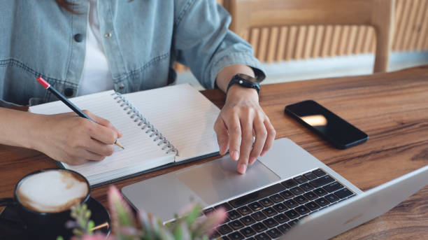 istock Woman sitting at table, writing on notebook, searching the information on laptop computer at coffee shop, closeup. Business woman taking note, planning work project on notebook, student studying online, e-learning 2166122699