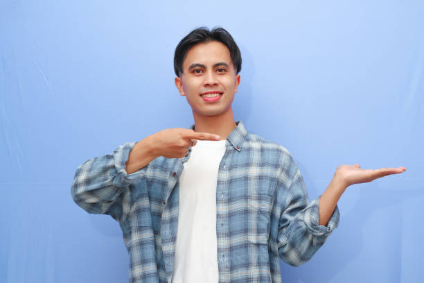 istock Attractive Asian man smiling while pointing to his open hand palm isolated on blue background 2166126017