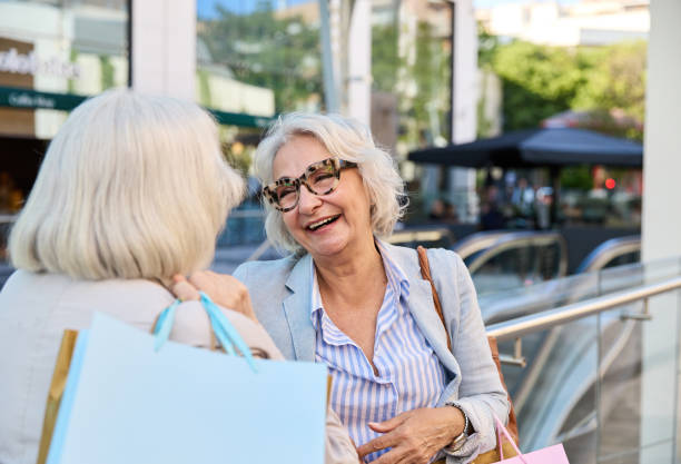 istock Smiling senior female speaking with unrecognizable friend with back view and looking at each other while standing together near metal railing with shopping mall paper bags in daylight 2166182493